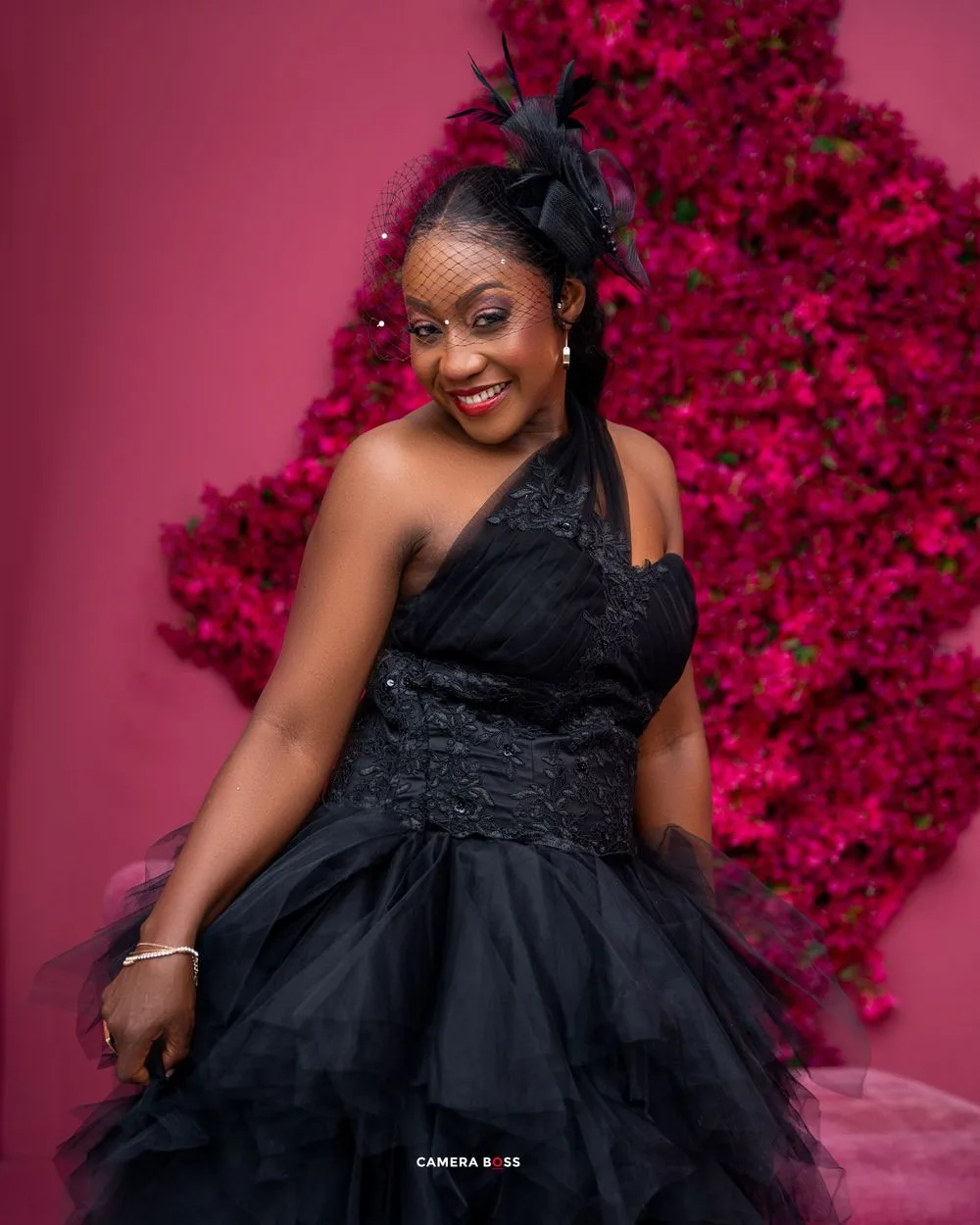 A glamorous portrait against a pink floral backdrop showing an elegant black one-shoulder tulle gown.