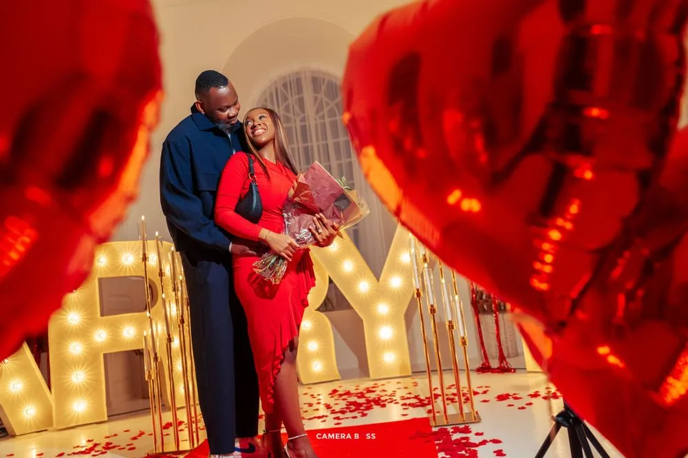 Romantic Valentine's Day proposal scene with lit marquee letters, red heart balloons and rose petals on the ground.