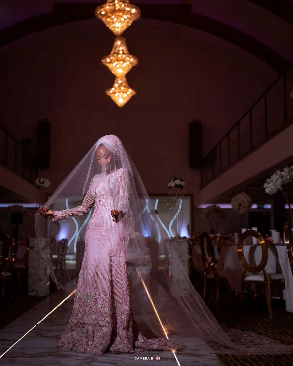 A bride in a blush pink lehenga gown poses on a runway stage with elegant chandeliers overhead.