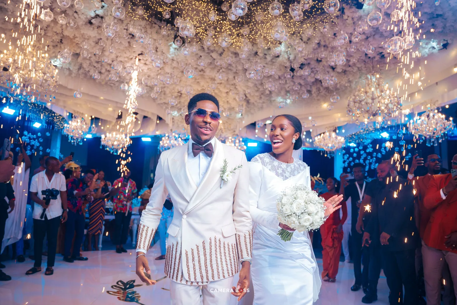 A couple in matching white attire celebrate at their wedding reception under a sparkling chandelier ceiling.