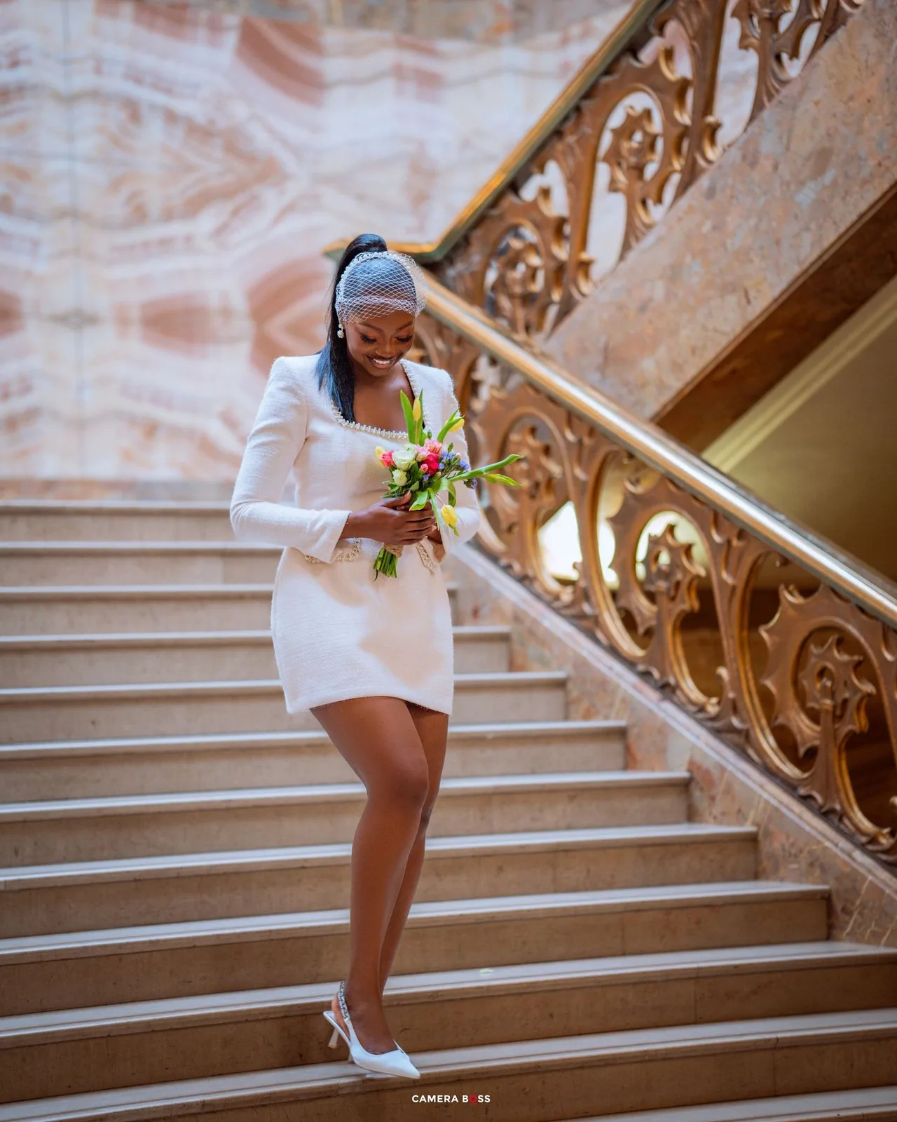 Person in white formal dress ascending ornate staircase while holding bouquet.