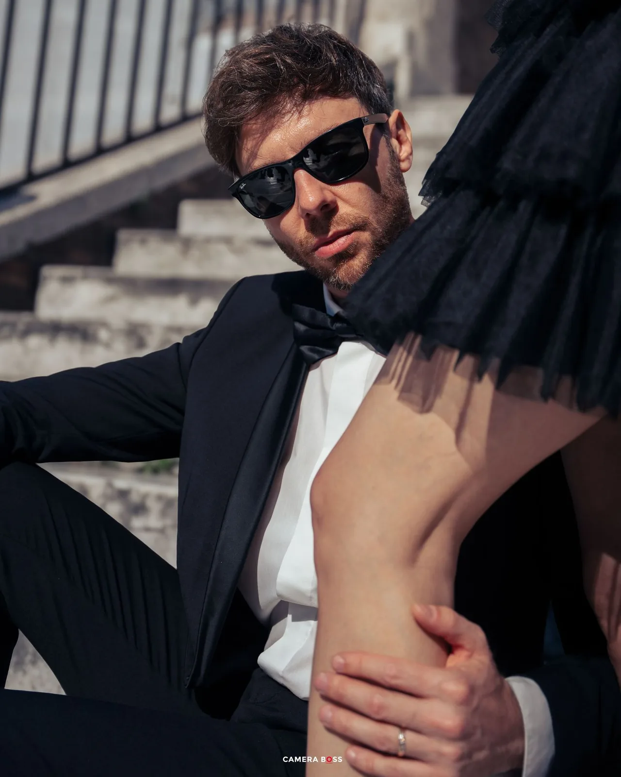 A person in a black tuxedo and sunglasses sits on stone steps, photographed in dramatic lighting.