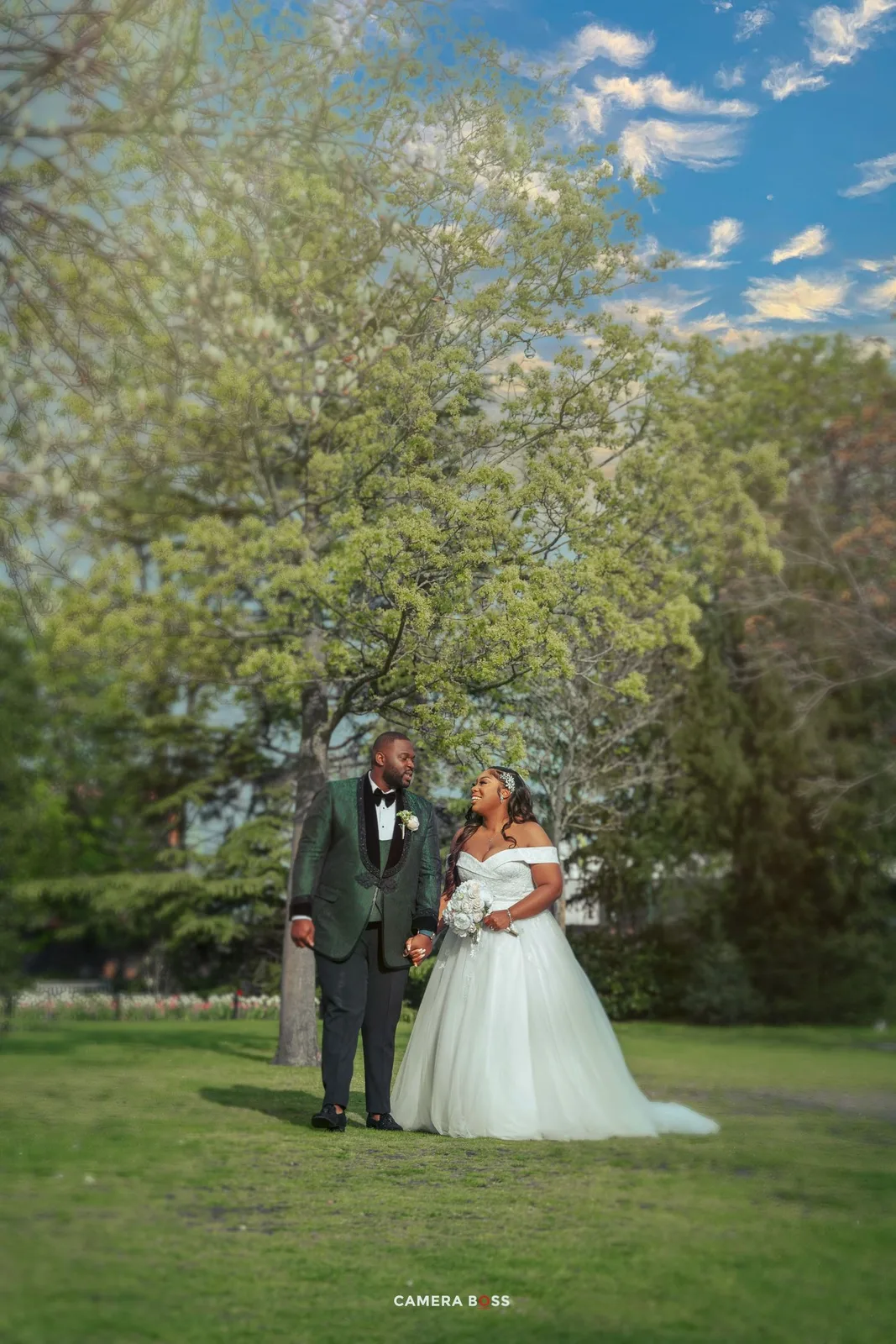 A couple shares a joyous moment in their wedding attire on a lush green lawn surrounded by blooming trees.