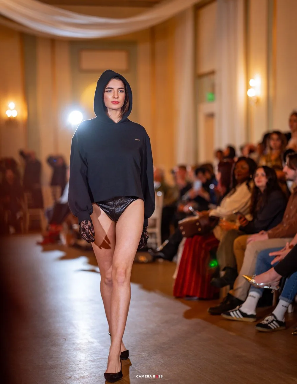 Model walks runway in a black hoodie and bodysuit at an indoor fashion show with audience members seated along sides.