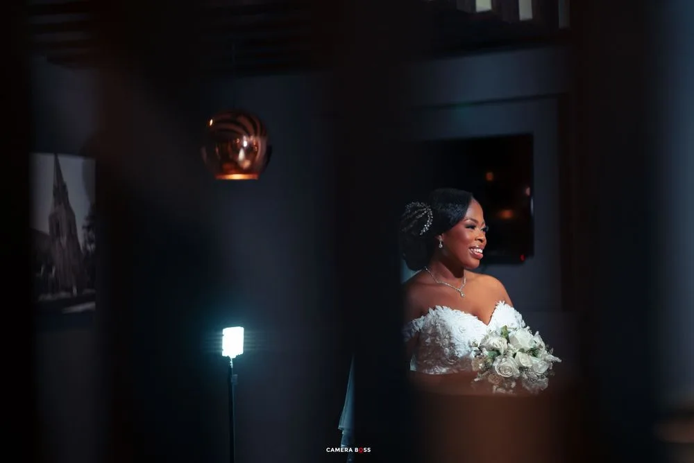 Bride in off-shoulder gown holds white bouquet while standing in dimly lit room with warm accent lighting.