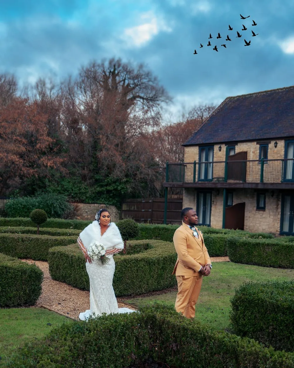 Two people in contrasting outfits standing in a manicured garden with hedges and a Tudor-style building in the background.