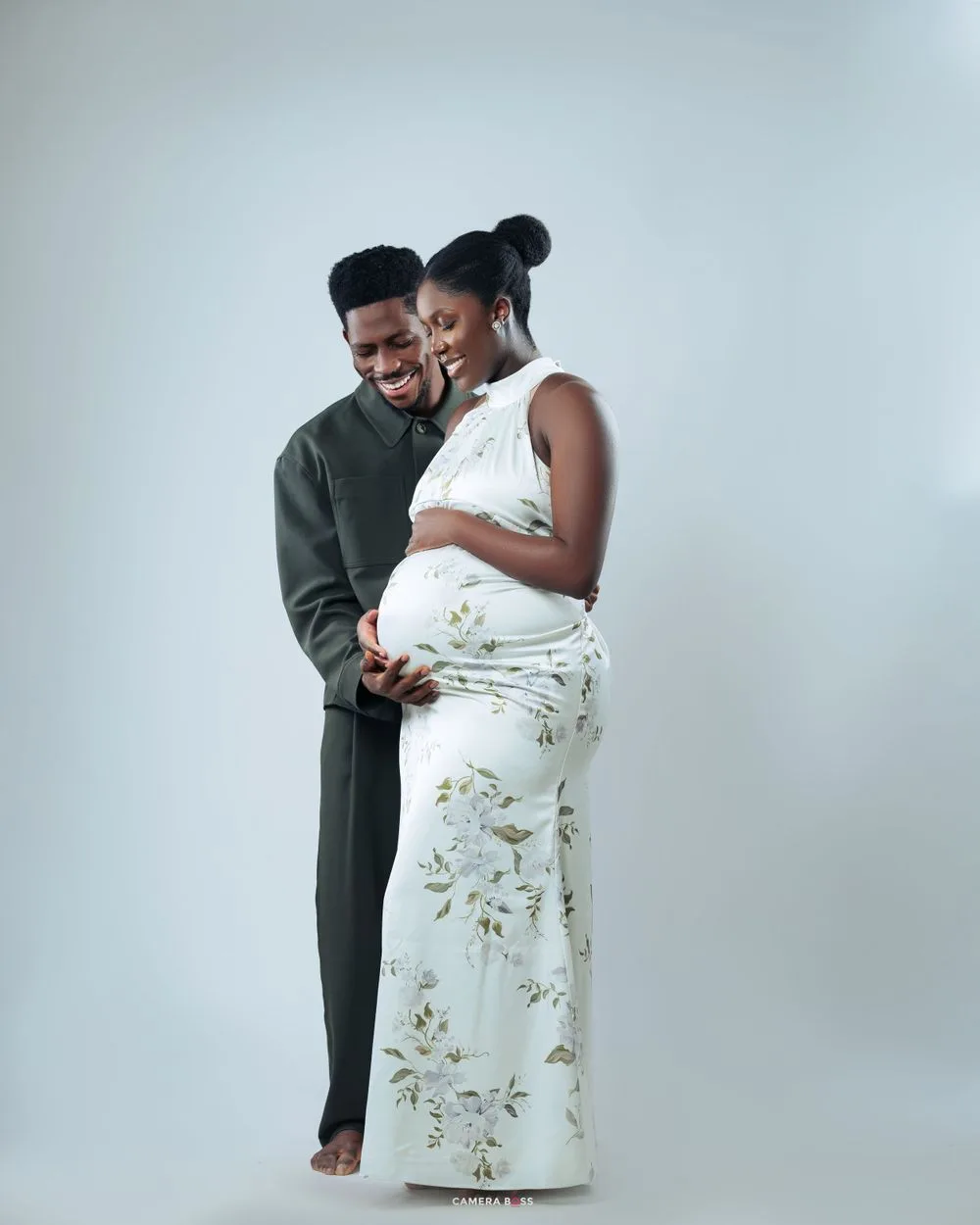 A couple shares a tender moment together against a light gray backdrop, the person wearing a floral white dress.