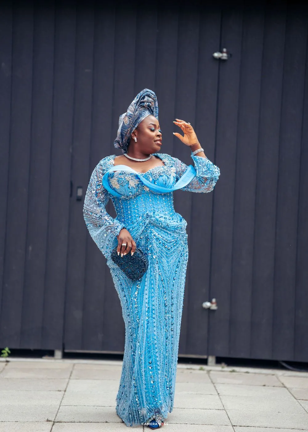 A person wearing an elegant turquoise blue sequined gown and matching head wrap poses dramatically against a dark wall.