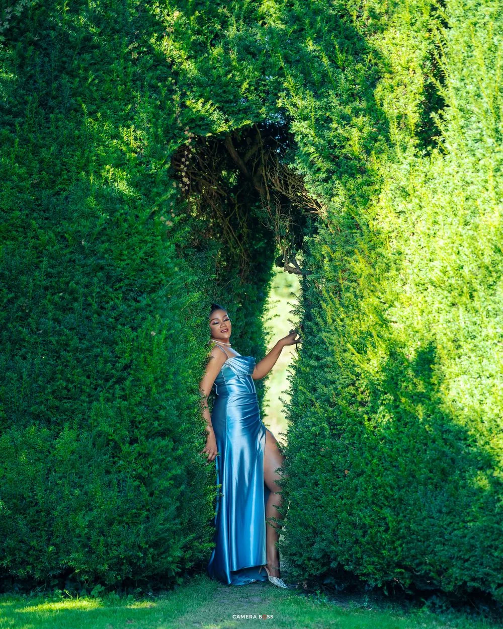 A figure in a flowing blue evening gown poses dramatically against a vivid green hedge in a sunlit garden setting.