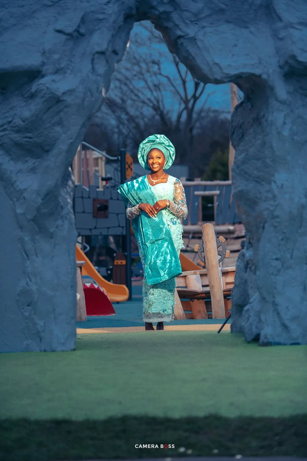 Figure in traditional turquoise Nigerian attire and gele headwrap stands framed by stone archway on outdoor stage set.