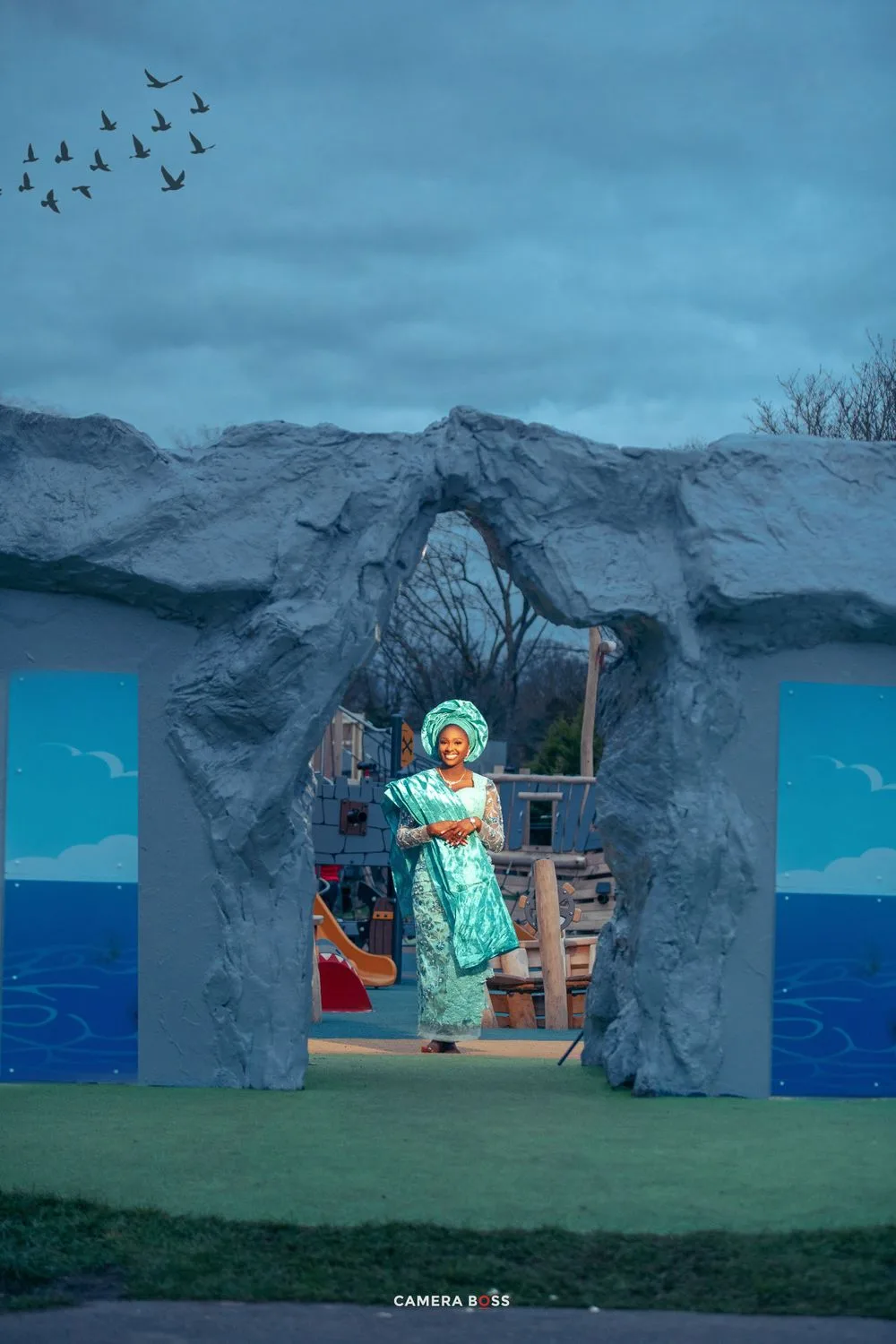 A stage set with stone archway and ocean backdrop features a performer in a turquoise dress holding a broom on the set.