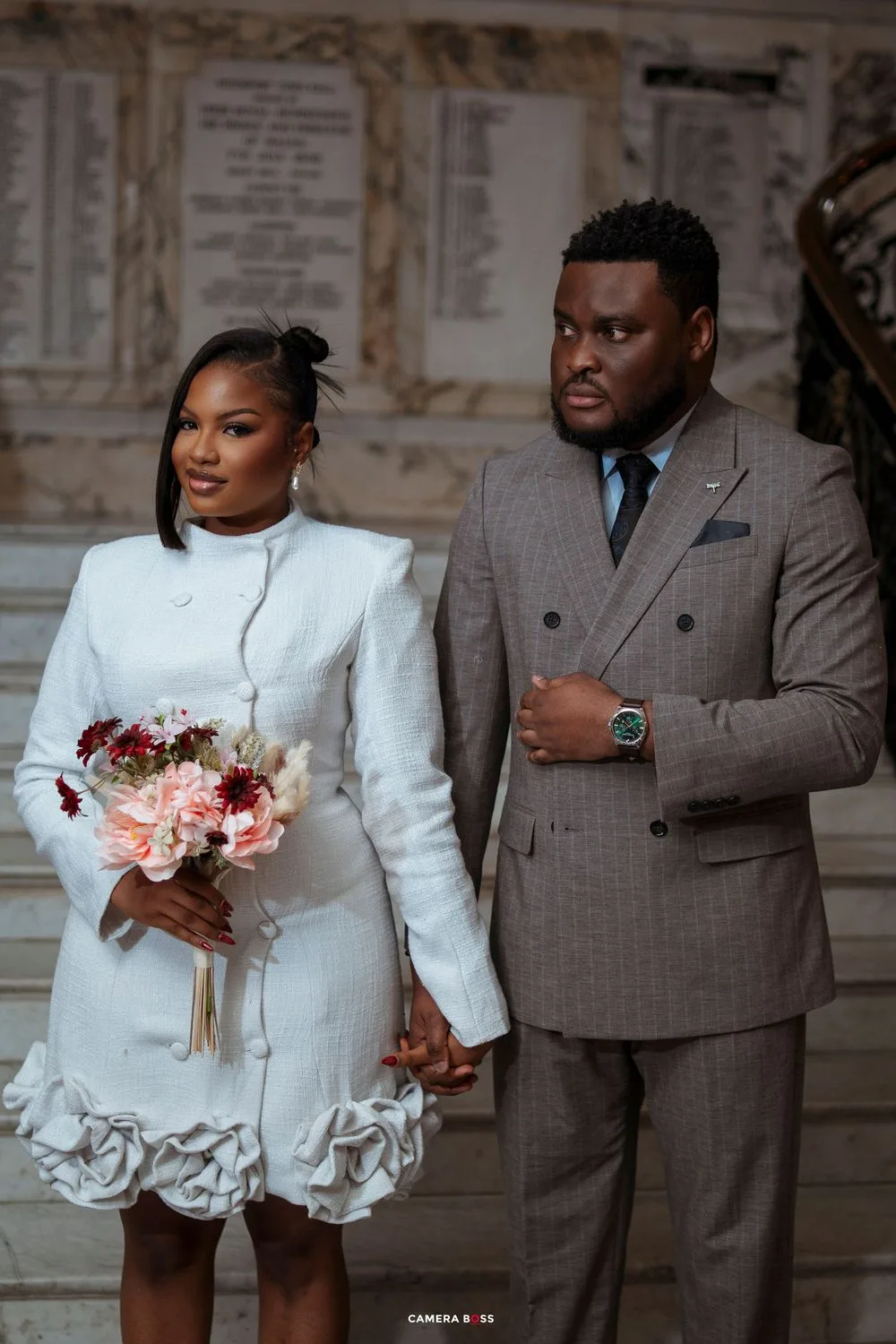 Elegant couple in wedding attire, bride in white dress with floral hem holding bouquet, groom in gray suit holding hands.