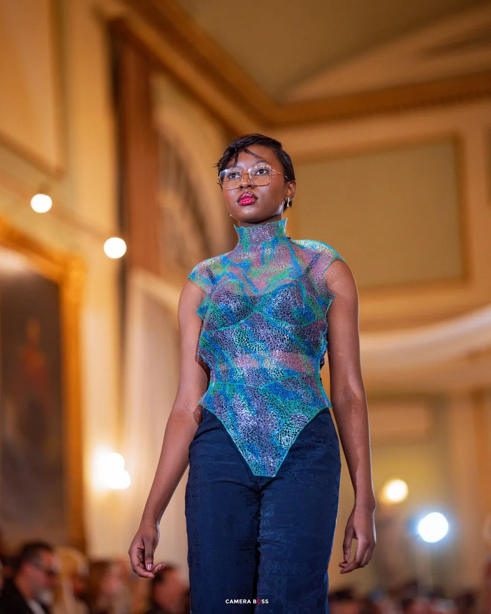 Fashion model wearing a shimmering blue and purple mesh bodysuit with jeans on an illuminated runway.