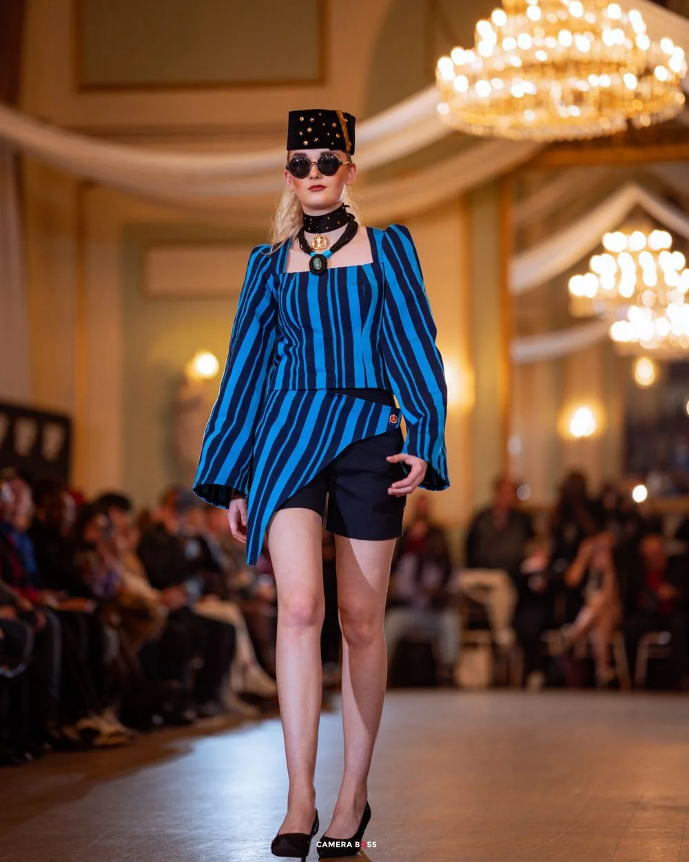 Fashion model walks runway in blue striped top and black shorts under elegant chandeliers at upscale fashion show.