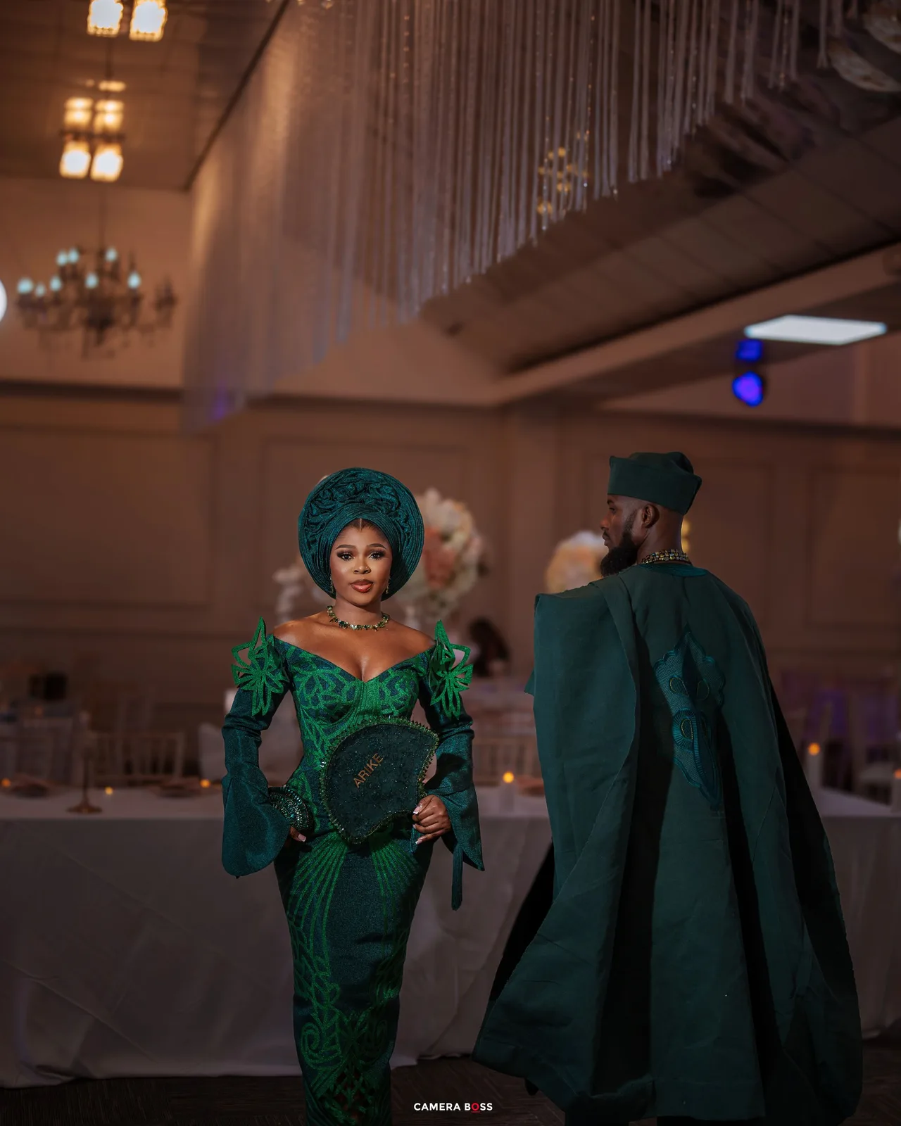 Two people in ornate emerald green traditional attire with matching headpieces at an elegant indoor venue.