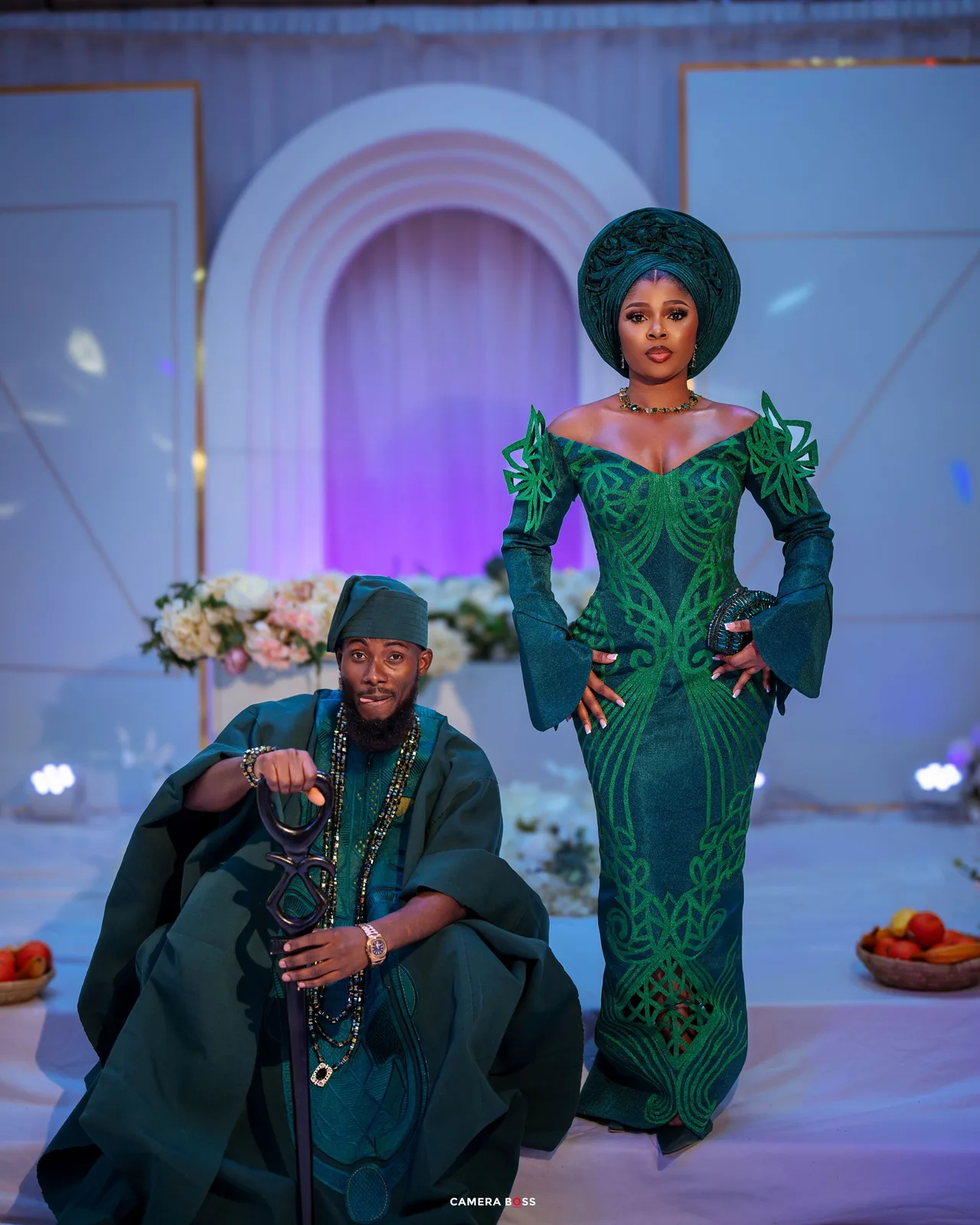 Two people in elegant emerald green traditional African attire pose together in front of an archway with purple lighting effects.