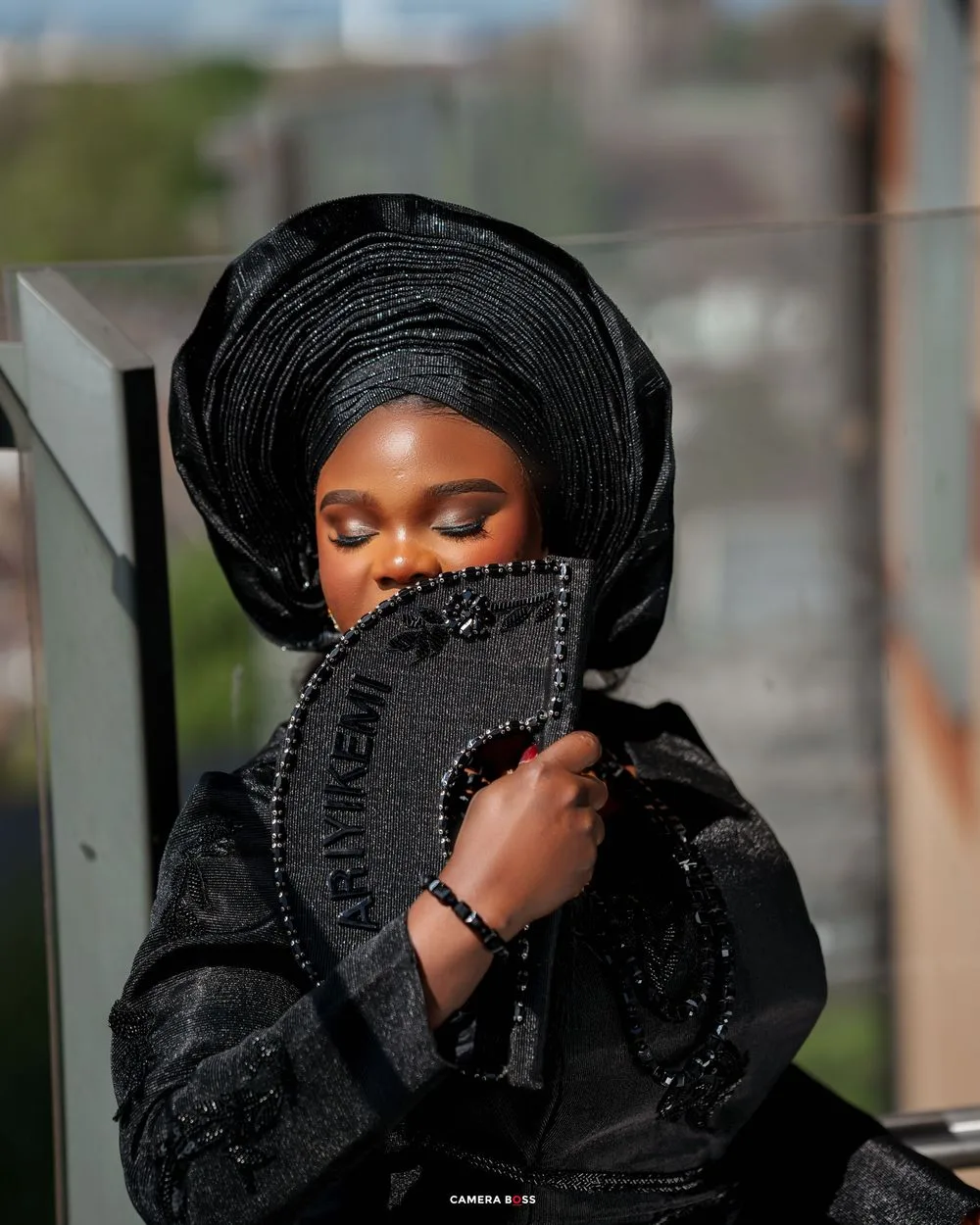 Person in traditional black African attire with gele headwrap poses with decorative black fan against blurred background.