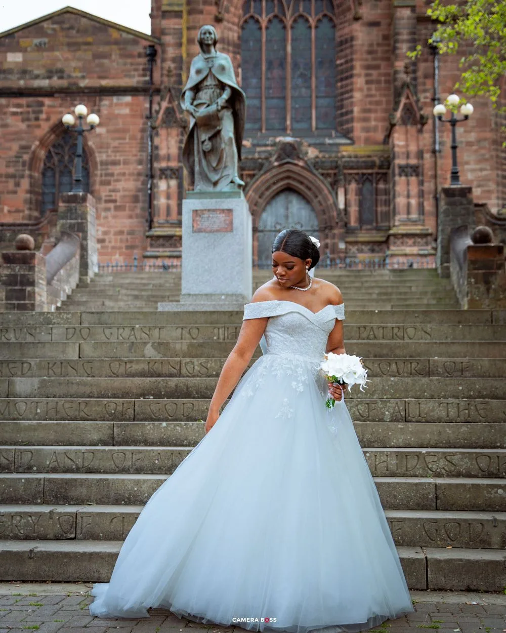 A bride in an elegant off-shoulder white ballgown poses on stone steps in front of a historic cathedral church.