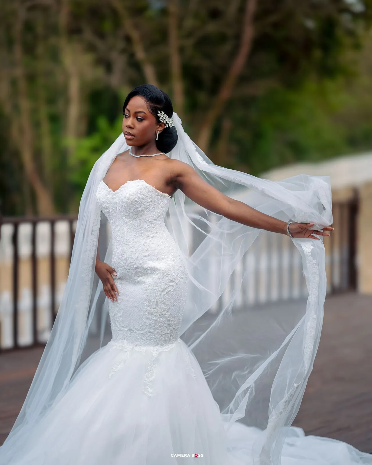 A bride in a stunning mermaid-style wedding gown with a flowing veil poses gracefully on a pathway surrounded by trees.