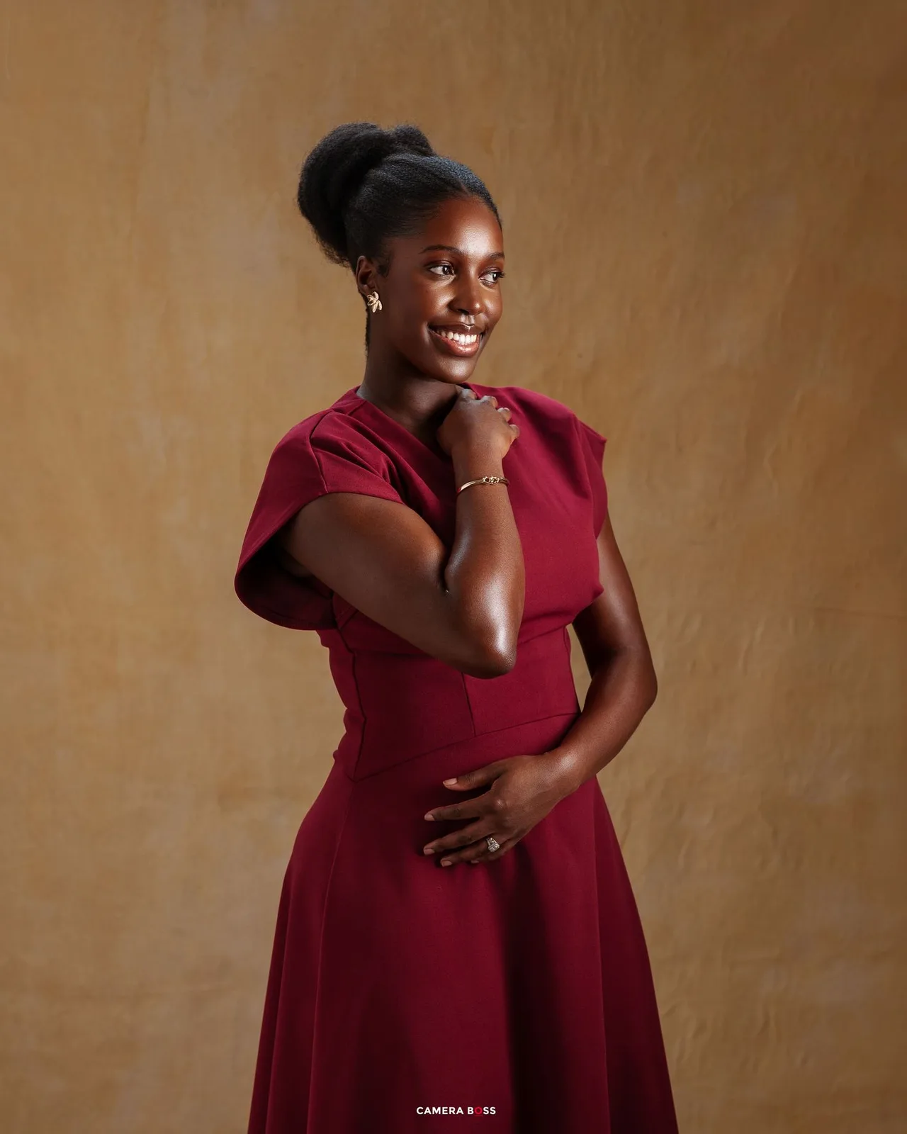 A person in a burgundy dress poses elegantly against a textured brown background with a warm smile.