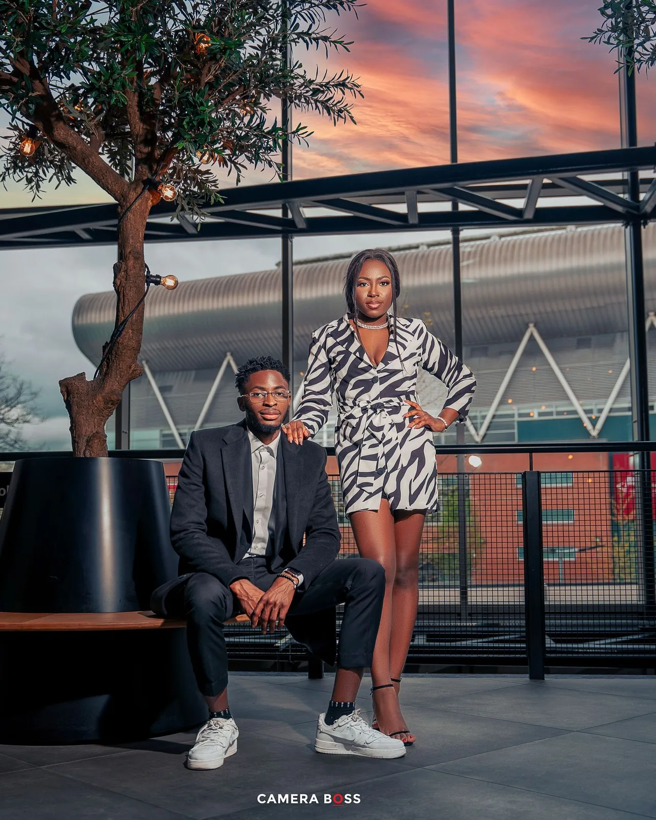 A stylish couple poses at sunset outside a modern building with dramatic orange sky and architectural details.