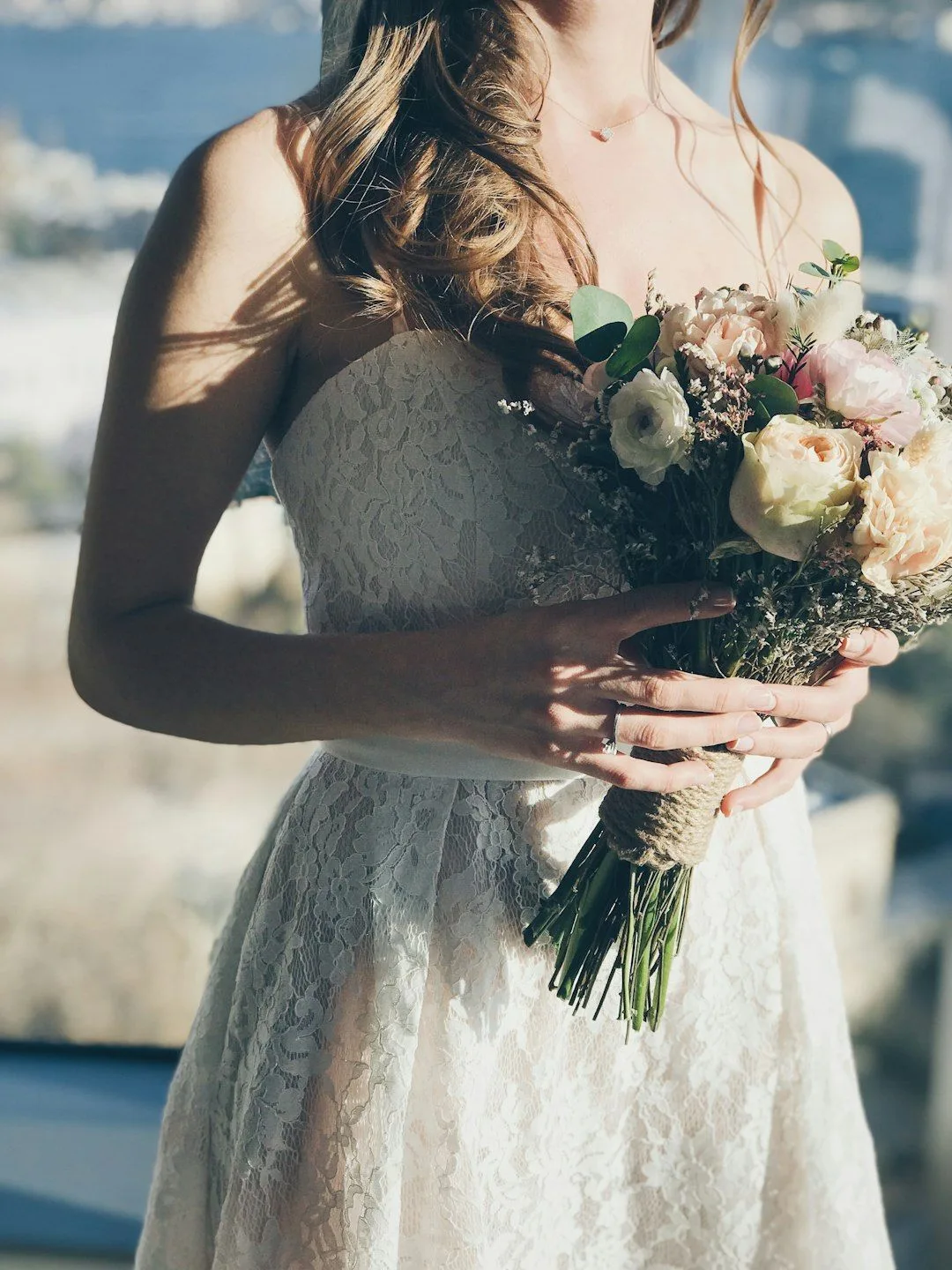 Close up of a bride in white lace dress holding a romantic bouquet with roses and mixed flowers against a blurred backdrop.