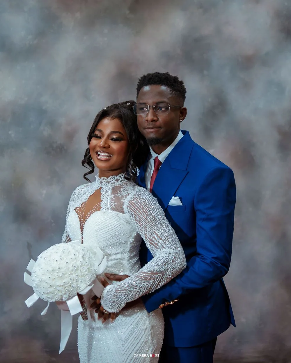 A couple in wedding attire pose together, with the bride in white lace and the groom in a royal blue suit.