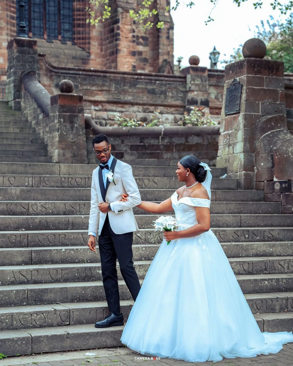 A newlywed couple in formal attire share a moment on ornate stone church steps during their wedding day.