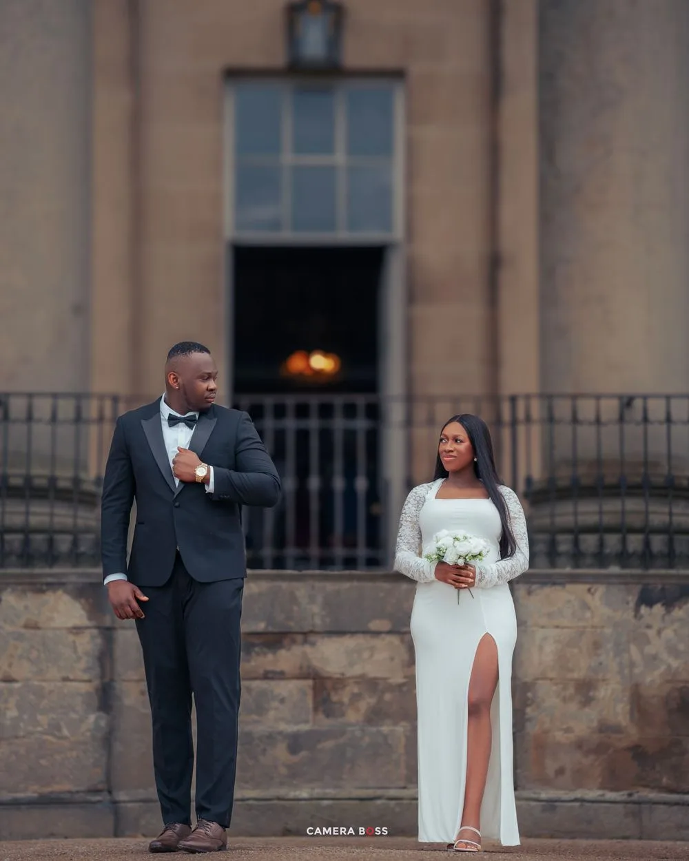 A bride and groom pose together outside a historic stone building during their wedding photoshoot at dusk.