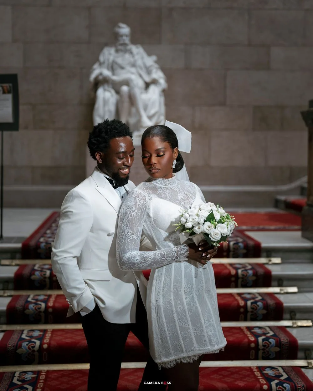 Elegant couple in white wedding attire pose before a grand statue in a formal hall, bride holding white bouquet.