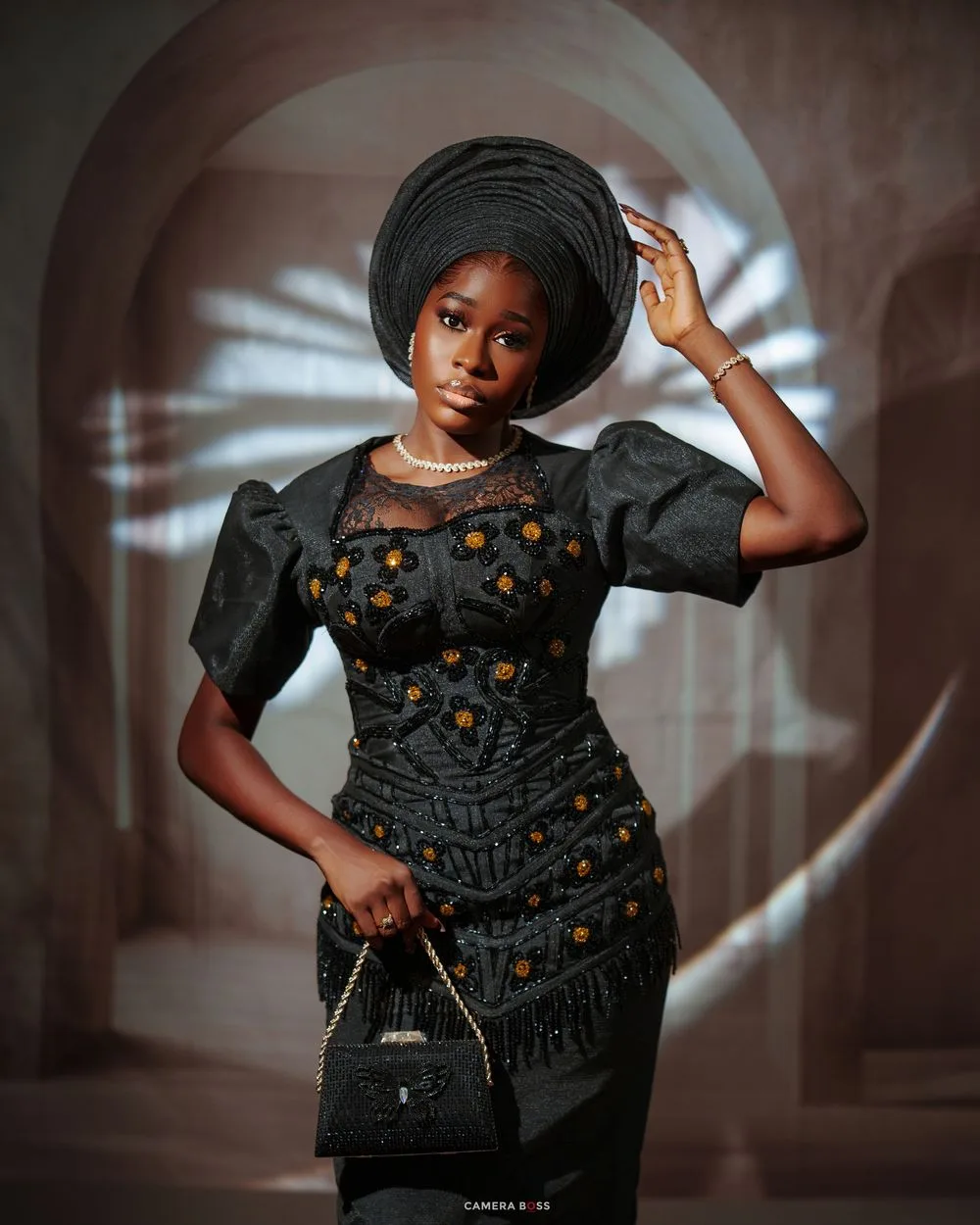 Woman in black embroidered dress and gele headwrap holding handbag in elegant Nigerian traditional attire.