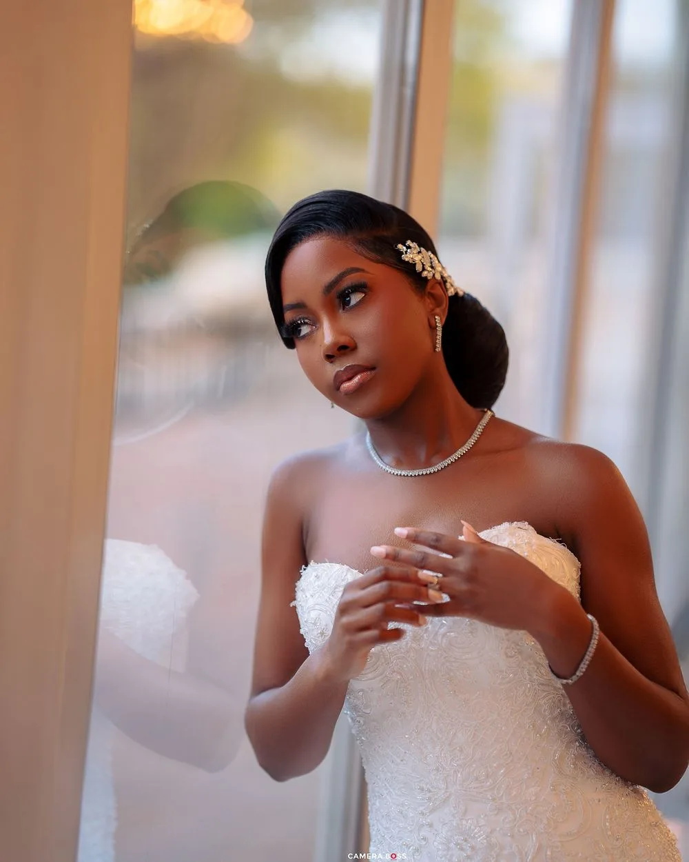 A bride in an elegant white strapless gown and pearl necklace poses thoughtfully by a sunlit window.