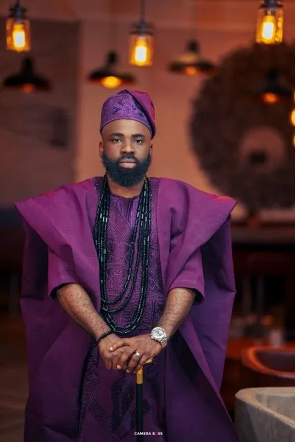Traditional African attire in rich purple with layered beaded necklaces and matching cap in atmospheric restaurant setting.