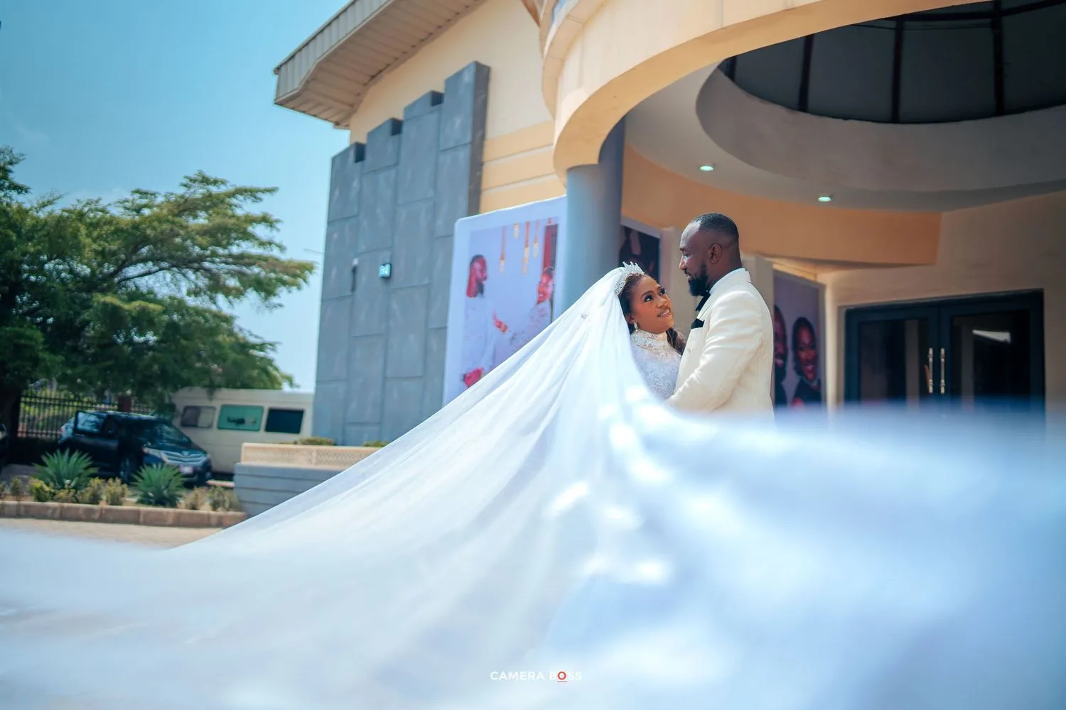 A couple in wedding attire share an intimate moment outside a building with the bride's long white veil flowing in the breeze.