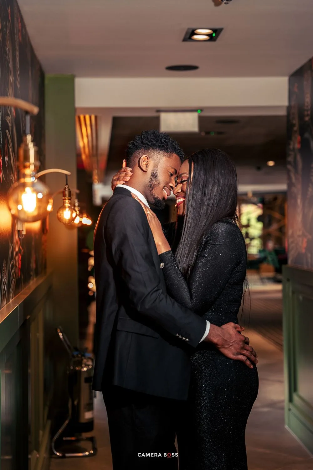 A romantic couple in elegant black attire share an intimate moment in a dimly lit hallway with warm ambient lighting.