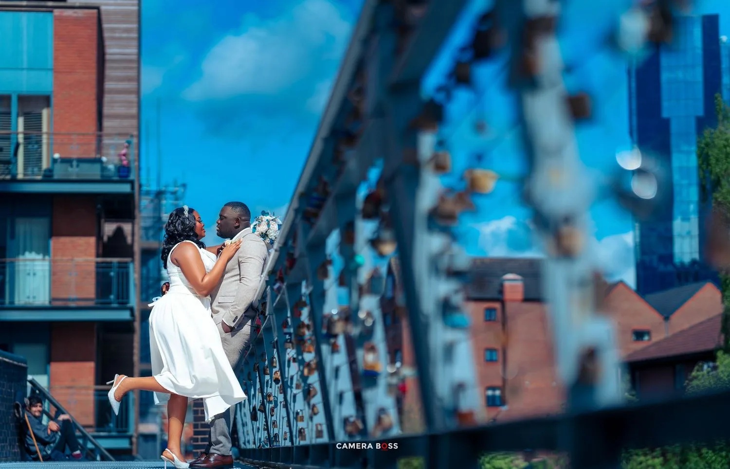A romantic couple embraces on an urban bridge with red brick buildings and blue sky in the background.