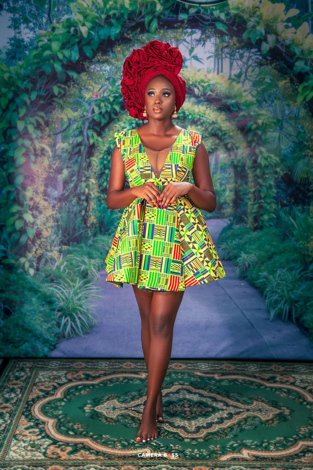 Woman in vibrant African print mini dress and red headwrap poses in garden setting.