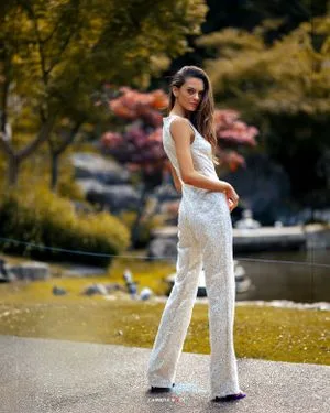 An elegant white lace jumpsuit is showcased in an outdoor setting with fall foliage and water in the background.