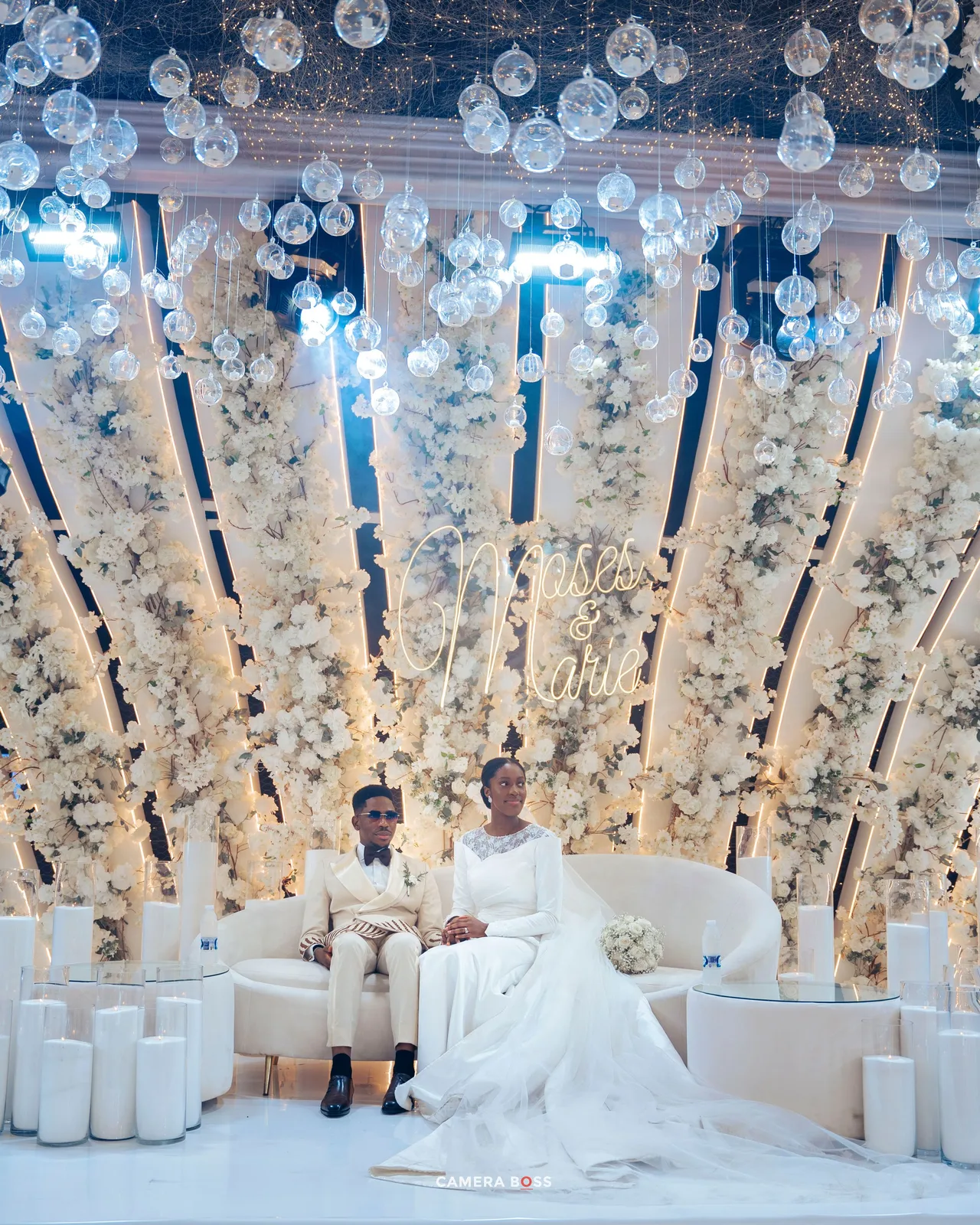 An elegant wedding ceremony setup with hanging glass orbs, white pillars and floral decorations against a light backdrop.