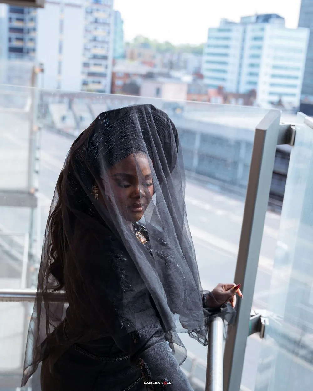 Person wearing black dress and veil poses on urban balcony with city buildings visible in background.