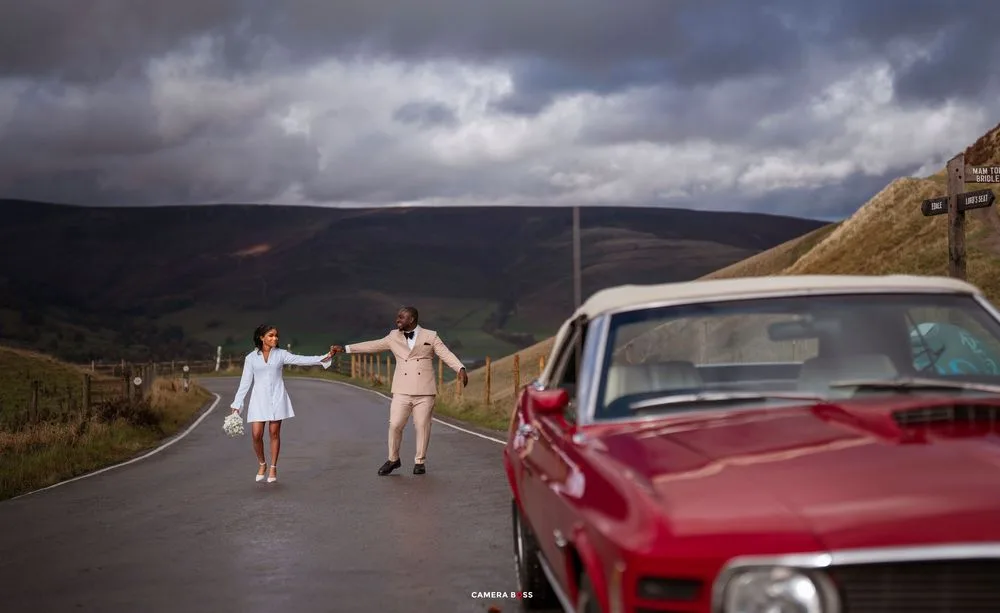Couple holding hands on a scenic road with rolling hills, dramatic clouds, and a classic red car in the foreground.