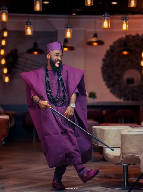 Traditional Nigerian attire in rich purple with matching cap and beads decorates a stylish ensemble in a warmly lit interior.