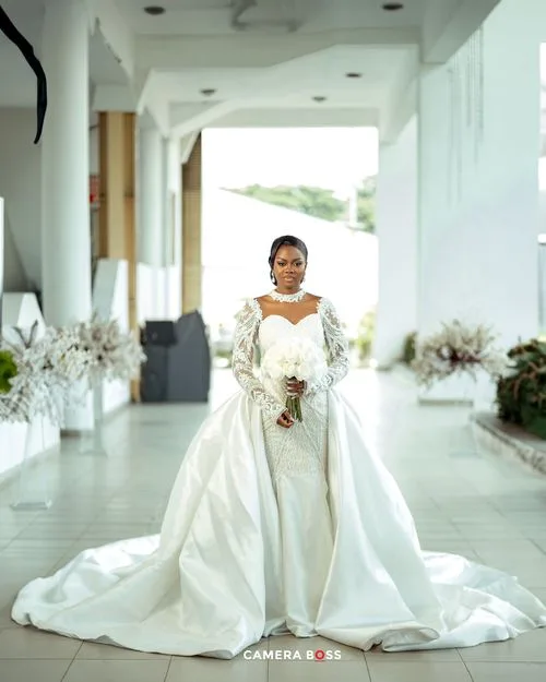 A bride poses in an elegant white wedding gown with long sleeves and dramatic train in a bright hallway.