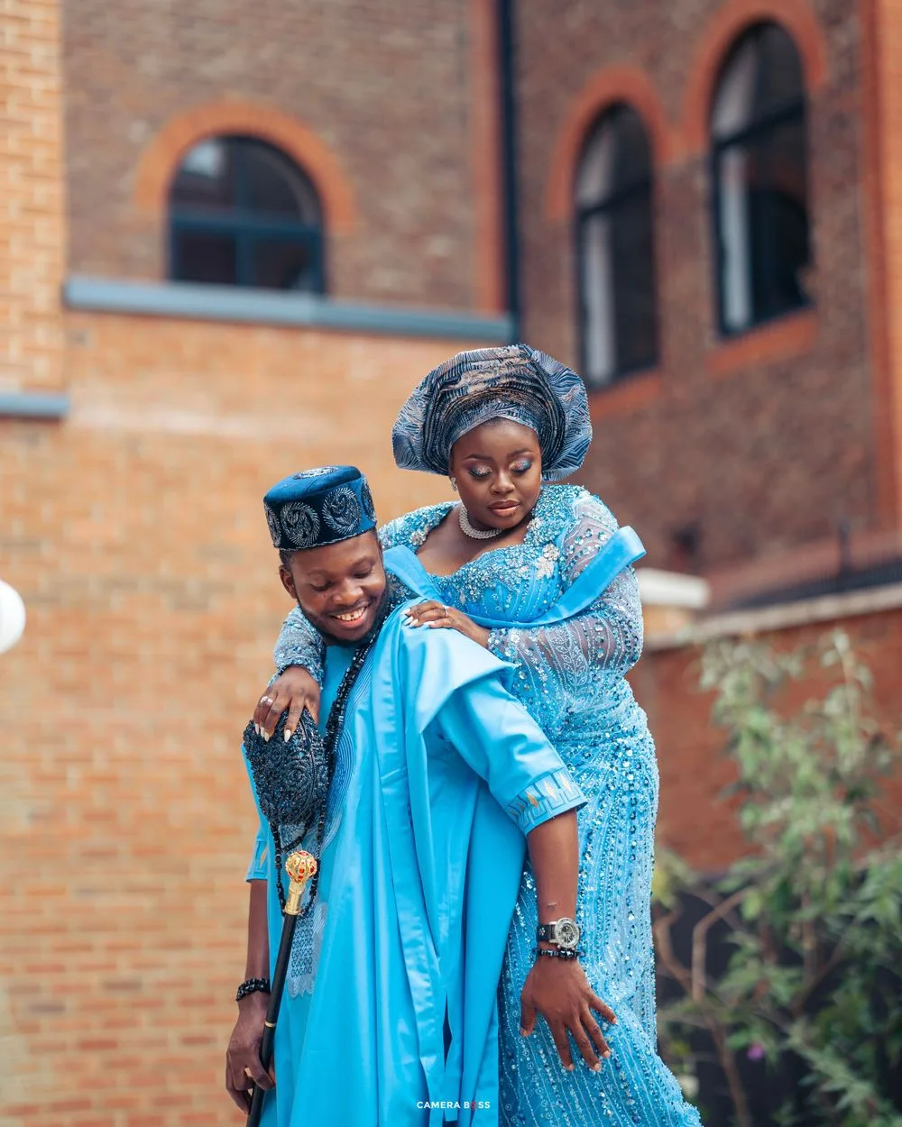Two people wearing vibrant blue traditional African attire embrace and smile outside a brick building.
