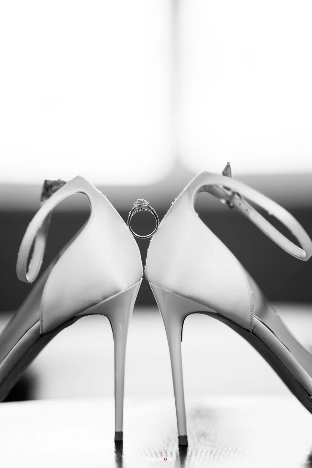 Black and white photo of elegant white high heels with a wedding ring nestled between them creating artistic symmetry.