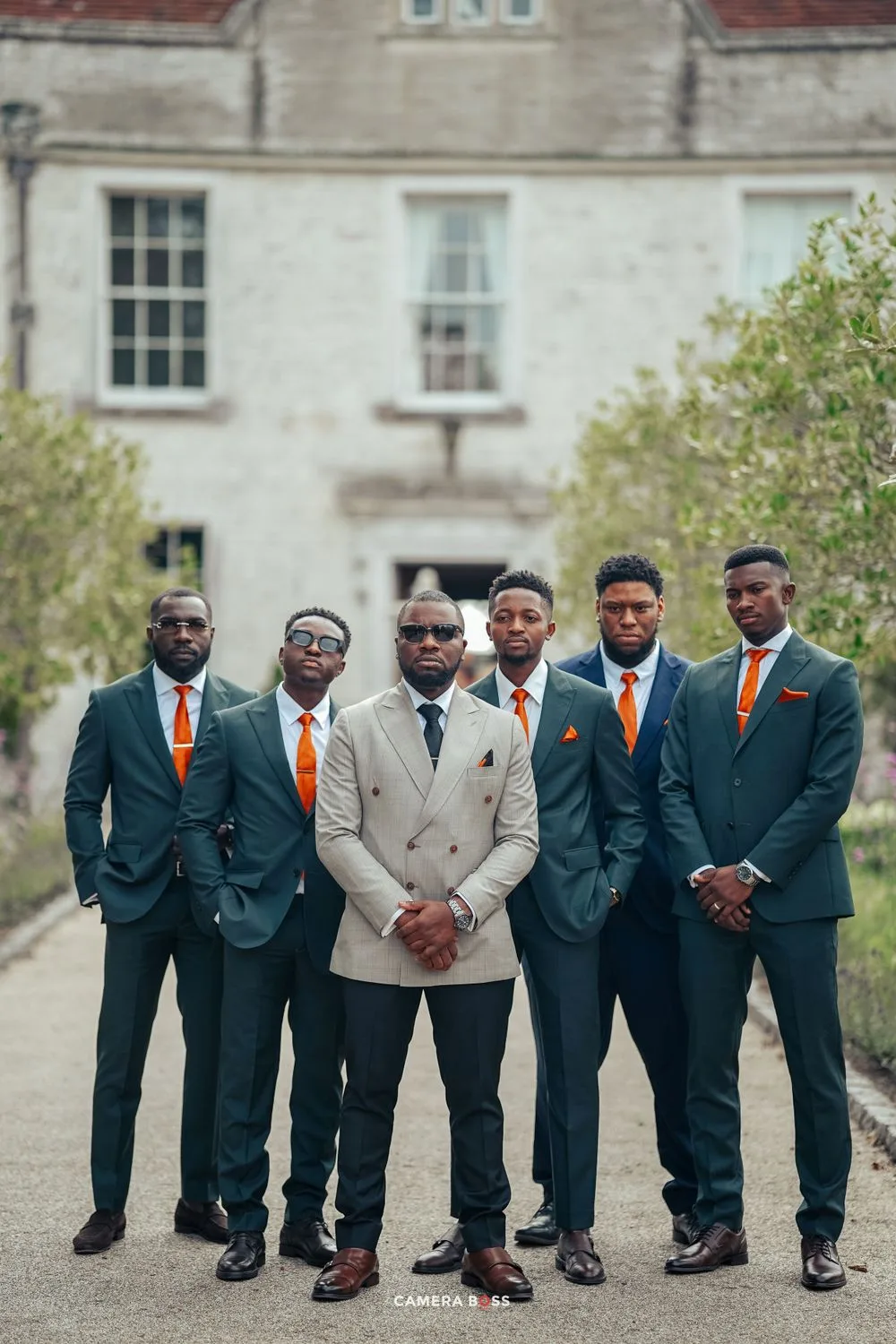 Wedding party in coordinated dark suits and orange ties pose together outside a historic building.