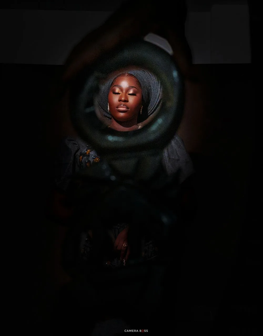 A young Black woman in a head wrap poses serenely, framed by dark hands forming a circle around her illuminated face.