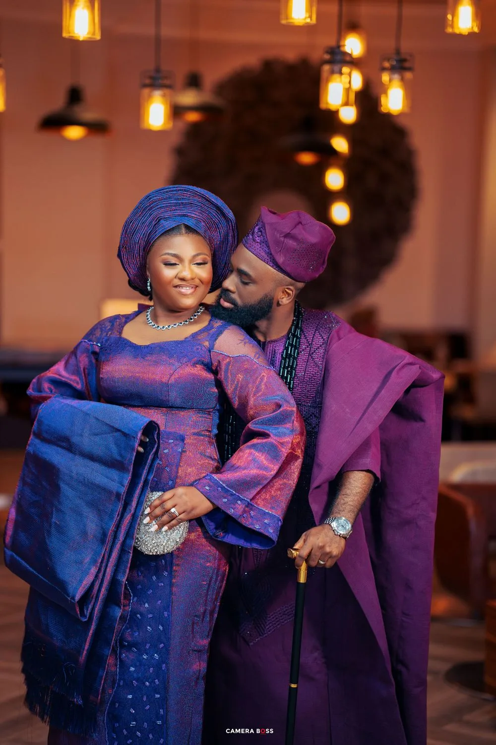 A couple in traditional African attire featuring blue and purple colors embrace under warm lighting with pendant lamps.