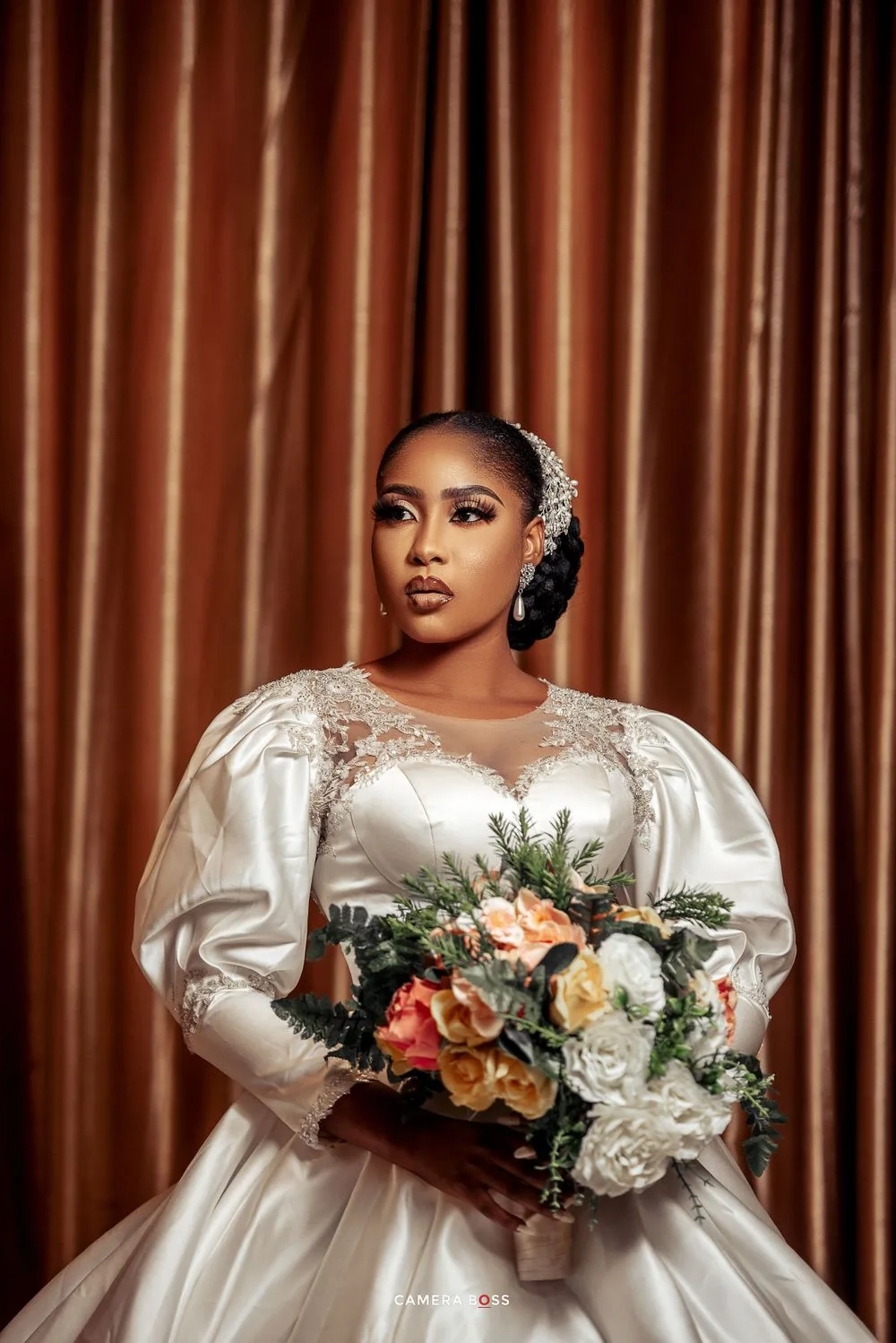Elegant bride in satin ball gown holds colorful bouquet against bronze curtain backdrop.