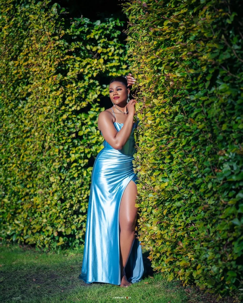 A figure in a flowing light blue satin evening gown poses elegantly against a lush green hedge backdrop.